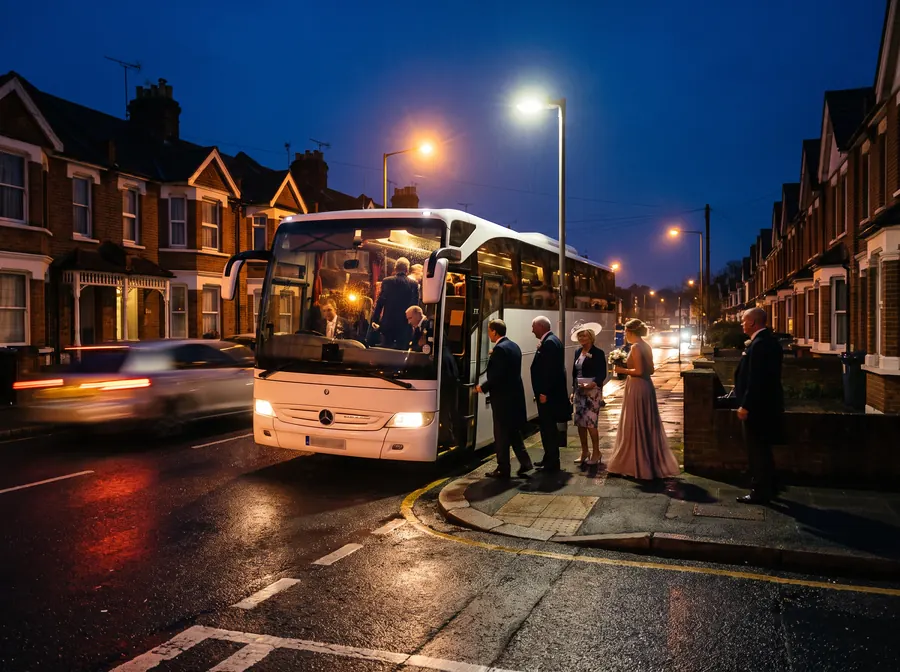 A white coach at night on a street of Epsom terraces, a wedding party in formal dress boarding under streetlights